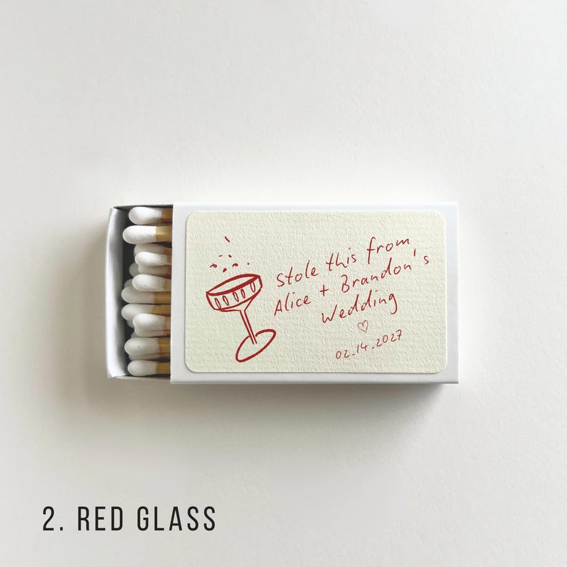May include: A matchbox with a cream-colored label featuring a red illustration of a champagne glass and the text "Stole this from Alice + Brandon's Wedding 02.14.2027". The match heads are white with wooden stems.