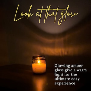 May include: A lit candle in a glass jar. The candle's flame casts a warm glow, with the text "Look at that glow" in cursive script at the top. The text "Glowing amber glass give a warm light for the ultimate cozy experience" is on the right.