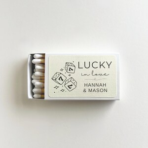 Könnte beinhalten: Eine weiße Streichholzschachtel mit einem cremefarbenen Etikett. Das Etikett zeigt drei Würfel mit der Zahl sieben, die Worte "LUCKY in love" und die Namen "HANNAH & MASON" in schwarzer Schrift.