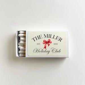 May include: A rectangular matchbox with the text "THE MILLER Holiday Club" and a red bow illustration. The matchbox contains white-tipped matches. The text "EST. 2014" is also present on the matchbox.