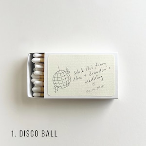 May include: A white matchbox with a cream-colored label featuring a disco ball illustration and handwritten text: "Stole this from Alice + Brandon's Wedding 02.14.2027". The match heads are white with a yellow tip.