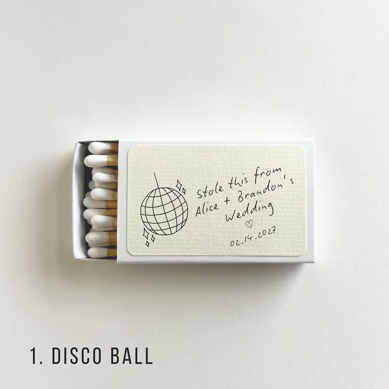 May include: A white matchbox with a cream-colored label featuring a disco ball illustration and handwritten text: "Stole this from Alice + Brandon's Wedding 02.14.2027". The match heads are white with a yellow tip.