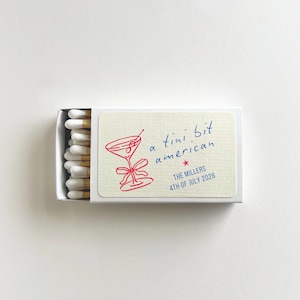 May include: A matchbox with a cream-colored label featuring a red cocktail illustration and the phrase "a tini bit american" in blue script. The text "THE MILLERS 4TH OF JULY 2026" is also included.