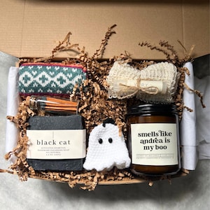 May include: A gift box filled with various items, including a black soap bar labeled "black cat", a candle that says "smells like andrea is my boo", a white ghost, a patterned sock, matches, and a bath sponge.