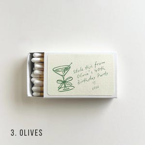 K&ouml;nnte beinhalten: Eine offene Streichholzschachtel mit einer gr&uuml;nen Illustration eines Martiniglases und dem Text "Stole this from Olivia's 40th Birthday Party" mit einem Herz und dem Jahr 2026. Enth&auml;lt wei&szlig;e Streichh&ouml;lzer mit gelben Spitzen.