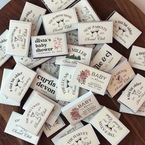 May include: A collection of personalized matchboxes with various text designs. The matchboxes feature text such as "New Parents," "Baby in Bloom," and names with dates. The color scheme is primarily white and cream with black text and illustrations.
