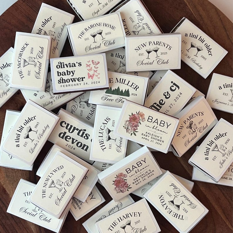 May include: A collection of personalized matchboxes with various text designs. The matchboxes feature text such as "New Parents," "Baby in Bloom," and names with dates. The color scheme is primarily white and cream with black text and illustrations.