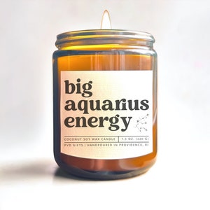 May include: A lit candle in a clear glass jar with a white label. The label reads "big aquarius energy" in black text, with the Aquarius constellation. The candle is 7.5 oz (220 g) and made with coconut soy wax.