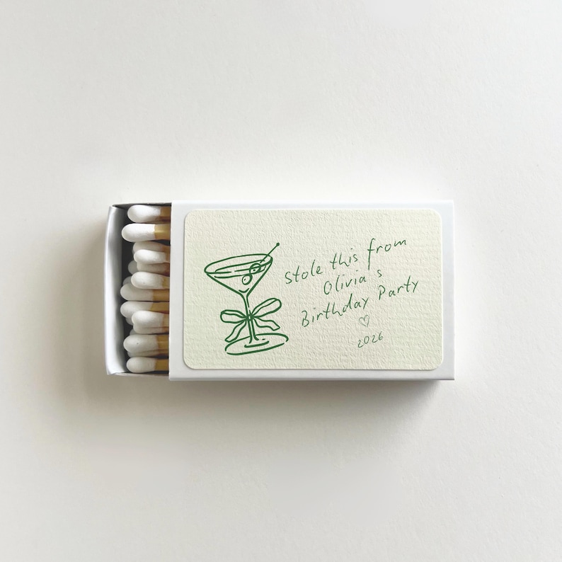Pu&ograve; includere: Una scatola di fiammiferi bianca con un'illustrazione verde di un bicchiere da martini e il testo "Stole this from Olivia's Birthday Party 2026". Le teste dei fiammiferi sono bianche con punte marroni.