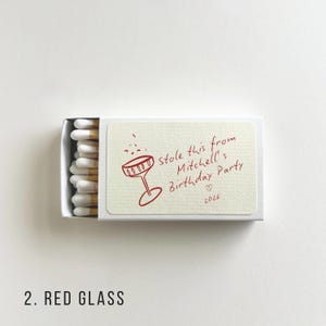 Pu&ograve; includere: Una scatola di fiammiferi bianca con un'illustrazione rossa di un bicchiere da cocktail e il testo "Stole this from Mitchell's Birthday Party 2026". La scatola contiene fiammiferi bianchi con punte gialle.