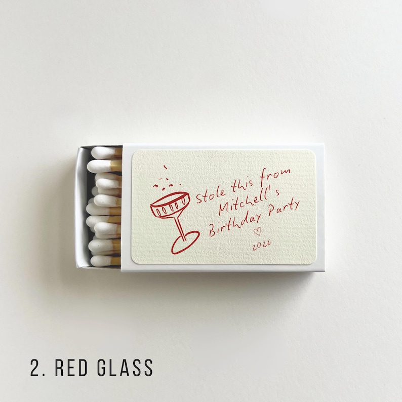 Pu&ograve; includere: Una scatola di fiammiferi bianca con un'illustrazione rossa di un bicchiere da cocktail e il testo "Stole this from Mitchell's Birthday Party 2026". La scatola contiene fiammiferi bianchi con punte gialle.