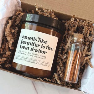 Könnte beinhalten: Ein braunes Kerzenglas mit schwarzem Deckel und einem weißen Etikett mit der Aufschrift "smells like jennifer is the best realtor" sowie ein kleines, durchsichtiges Glasfläschchen mit Holzstreichhölzern, verpackt in einem Karton mit Papierfüllung.