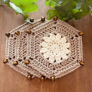 Può includere: Set di quattro sottobicchieri esagonali all'uncinetto marroni con un fiore all'uncinetto bianco al centro di ogni sottobicchiere. Ogni sottobicchiere ha perline di legno attorno ai bordi.