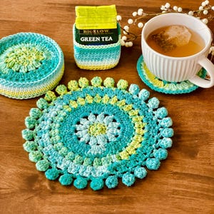May include: Handmade crochet tea set in shades of teal, yellow, and white. Includes a round coaster, a tea bag holder, and a matching coaster under a white teacup. The tea bag holder has "GREEN TEA" printed on it.