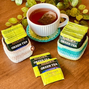 May include: A white teacup with tea and a tea bag sits on a crocheted coaster. Two crocheted baskets hold Bigelow Green Tea packets. Two additional tea packets are also visible. The tea packets are yellow and green.