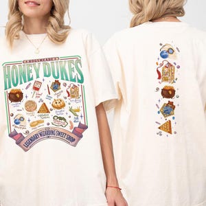 May include: Cream-colored t-shirt with a colorful graphic of the Honeydukes sweet shop. The front features the shop's name and various sweets. The back has a vertical arrangement of illustrated treats and a logo.