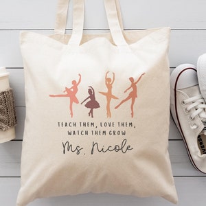 May include: A beige canvas tote bag with a design of five dancers in shades of pink and orange. The text "Teach them, love them, watch them grow Ms. Nicole" is printed on the bag.