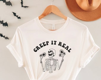 Puede incluir: Camiseta blanca con un esqueleto negro sosteniendo flores y el texto "Creep It Real" en negro.