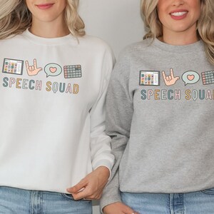 May include: Two sweatshirts, one white and one grey, display the text "SPEECH SQUAD" in various colours. The design includes a tablet, a hand in sign language, a speech bubble with a heart, and a keyboard.