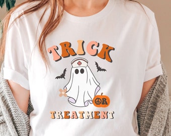 Pode incluir: Camiseta branca com um gráfico temático de Halloween. O design apresenta as palavras "Trick Treatment" em tons de laranja e pêssego, um fantasma usando um chapéu de enfermeira, morcegos e uma abóbora com um símbolo de paz.