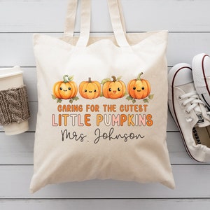 K&ouml;nnte beinhalten: Eine wei&szlig;e Stofftasche mit einem Design aus vier orangefarbenen K&uuml;rbissen mit l&auml;chelnden Gesichtern. Der Text auf der Tasche lautet "Caring for the cutest little pumpkins Mrs. Johnson".