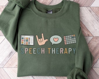 May include: A green sweatshirt with the text "SPEECH THERAPY" in colorful letters. The design includes a tablet with speech therapy symbols, a hand making the sign for "I love you", a speech bubble with a heart, and a grid of speech therapy symbols.