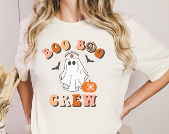 May include: White t-shirt with a graphic of a ghost wearing a nurse's hat and holding a pumpkin. The text "Boo Boo Crew" is written in a retro font with a peace sign.