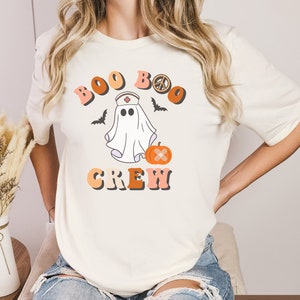 May include: White t-shirt with a graphic of a ghost wearing a nurse's hat and holding a pumpkin. The text "Boo Boo Crew" is written in a retro font with a peace sign.