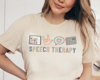 Può includere: Una maglietta beige con un design grafico che presenta un tablet, una mano che fa il segno 'Ti amo', un fumetto con un cuore e una griglia di quadrati. Il testo 'SPEECH THERAPY' è stampato sotto il grafico. La maglietta è indossata da una persona che indossa jeans blu. La parola 'NATURALE' è stampata in marrone su uno sfondo bianco.