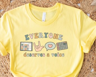 Puede incluir: Camiseta amarilla con el texto "Everyone deserves a voice" en letras de colores. El diseño incluye una mano haciendo el signo de "Te amo", una burbuja de diálogo con un corazón y un teclado.
