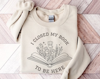 Può includere: Una felpa beige con la scritta "Ho chiuso il mio libro per essere qui" e un'immagine di un libro con dei fiori che ne escono.