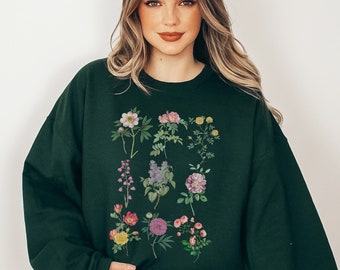 Könnte beinhalten: Ein dunkelgrüner Pullover mit einem Blumendruck, der verschiedene Blumen in den Farben Rosa, Lila, Gelb und Weiß zeigt. Die Blumen sind in einem Rastermuster auf der Vorderseite des Pullovers angeordnet.