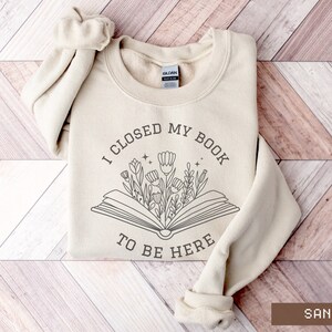 K&ouml;nnte beinhalten: Ein beiger Pullover mit dem Text "Ich habe mein Buch geschlossen, um hier zu sein" und einer Grafik eines Buches, aus dem Blumen wachsen.