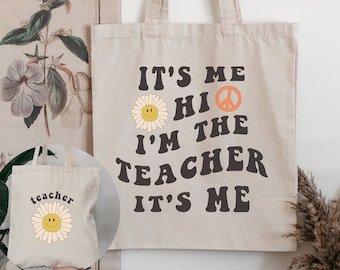 Può includere: Una borsa tote in tela beige con il testo "It's Me Hi I'm the Teacher It's Me" in nero. Sulla borsa sono presenti anche un segno di pace e una margherita gialla con un viso sorridente.