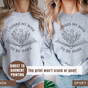 K&ouml;nnte beinhalten: Zwei graue Sweatshirts mit dem Text "Ich habe mein Buch geschlossen, um hier zu sein" darauf gedruckt. Der Text ist umgeben von einer Zeichnung eines offenen Buches, aus dem Blumen wachsen. Die Sweatshirts werden auf Models gezeigt, die Jeans tragen. Der Text "Direktdruck auf Kleidung" ist unten links im Bild gedruckt. Der Text "Der Druck rei&szlig;t nicht und l&ouml;st sich nicht ab!" ist unten rechts im Bild gedruckt. Der Text "Ash" ist unten links im Bild gedruckt. Der Text "Sport Grey" ist unten rechts im Bild gedruckt.