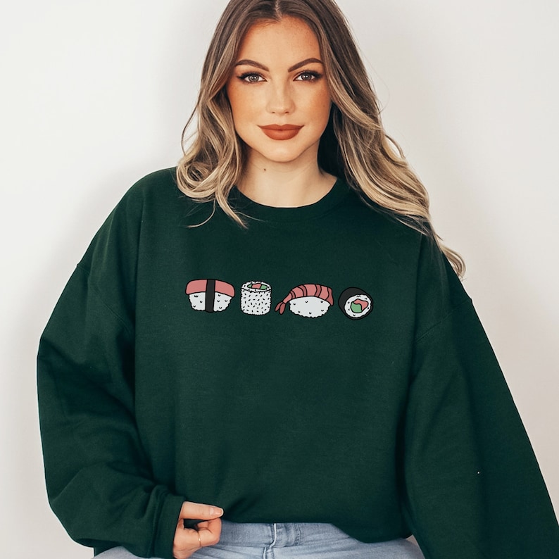 May include: A dark green sweatshirt with a graphic of five different sushi rolls. The rolls are drawn in a cartoon style and include a shrimp roll, a cucumber roll, a salmon roll, and two other rolls.