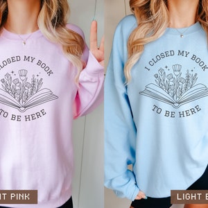 K&ouml;nnte beinhalten: Zwei Sweatshirts mit einem grafischen Design eines Buches, aus dem Blumen herauswachsen. Der Text auf den Sweatshirts lautet "Ich habe mein Buch geschlossen, um hier zu sein". Ein Sweatshirt ist hellrosa und das andere hellblau.