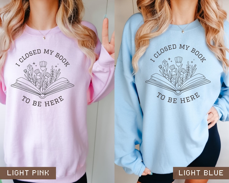 K&ouml;nnte beinhalten: Zwei Sweatshirts mit einem grafischen Design eines Buches, aus dem Blumen herauswachsen. Der Text auf den Sweatshirts lautet "Ich habe mein Buch geschlossen, um hier zu sein". Ein Sweatshirt ist hellrosa und das andere hellblau.