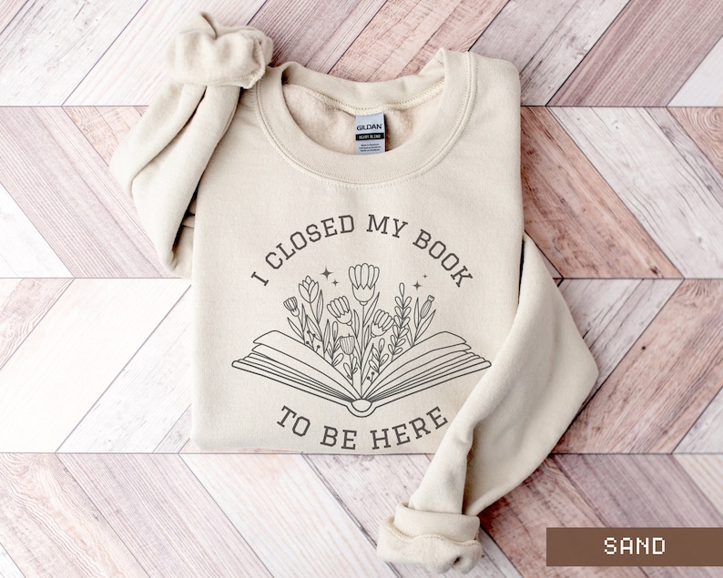 K&ouml;nnte beinhalten: Ein beiger Pullover mit dem Text "Ich habe mein Buch geschlossen, um hier zu sein" und einer Grafik eines Buches, aus dem Blumen wachsen.
