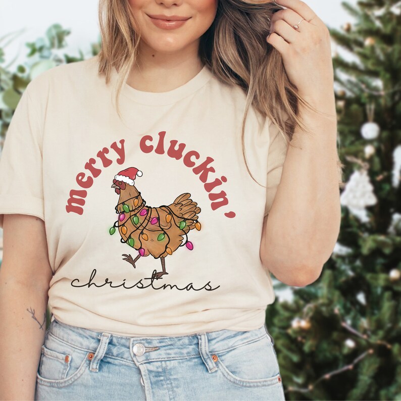 May include: A beige t-shirt with a red and white graphic of a chicken wearing a Santa hat and Christmas lights. The text "merry cluckin' christmas" is printed on the shirt.