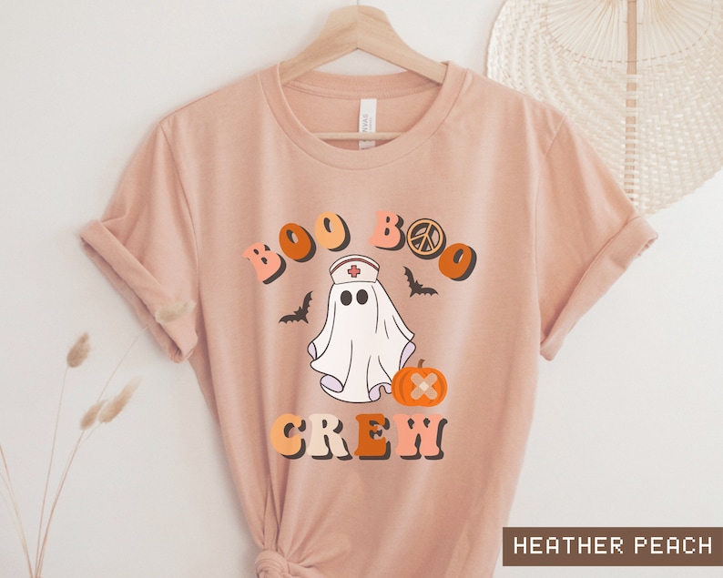 May include: A peach colored t-shirt with a graphic of a ghost wearing a nurse's hat and holding a pumpkin. The text "BOO BOO CREW" is above the ghost.