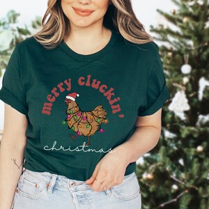 May include: A green t-shirt with a red and white design featuring a chicken wearing a Santa hat and Christmas lights. The text "merry cluckin' christmas" is printed on the shirt.
