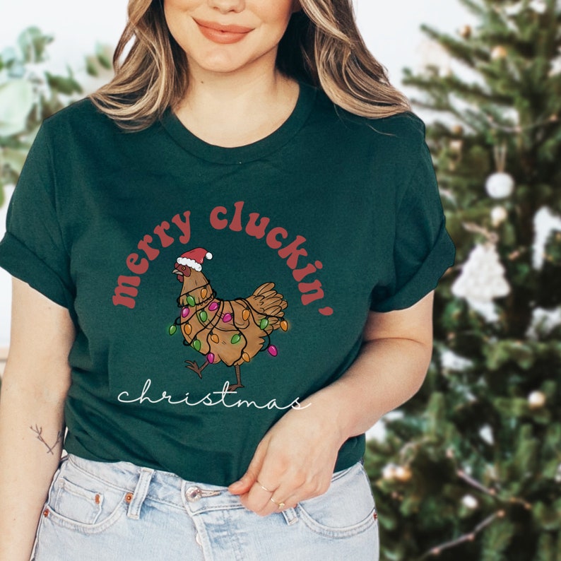 May include: A green t-shirt with a red and white design featuring a chicken wearing a Santa hat and Christmas lights. The text "merry cluckin' christmas" is printed on the shirt.