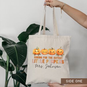 K&ouml;nnte beinhalten: Eine beige Tragetasche mit einem Design aus l&auml;chelnden orangefarbenen K&uuml;rbissen und dem Text "Caring for the cutest little pumpkins Mrs. Johnson" auf der Vorderseite. Die Tasche hat ein seitliches Etikett mit der Aufschrift "Side One".