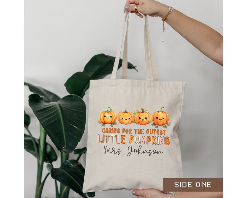 K&ouml;nnte beinhalten: Eine beige Tragetasche mit einem Design aus l&auml;chelnden orangefarbenen K&uuml;rbissen und dem Text "Caring for the cutest little pumpkins Mrs. Johnson" auf der Vorderseite. Die Tasche hat ein seitliches Etikett mit der Aufschrift "Side One".
