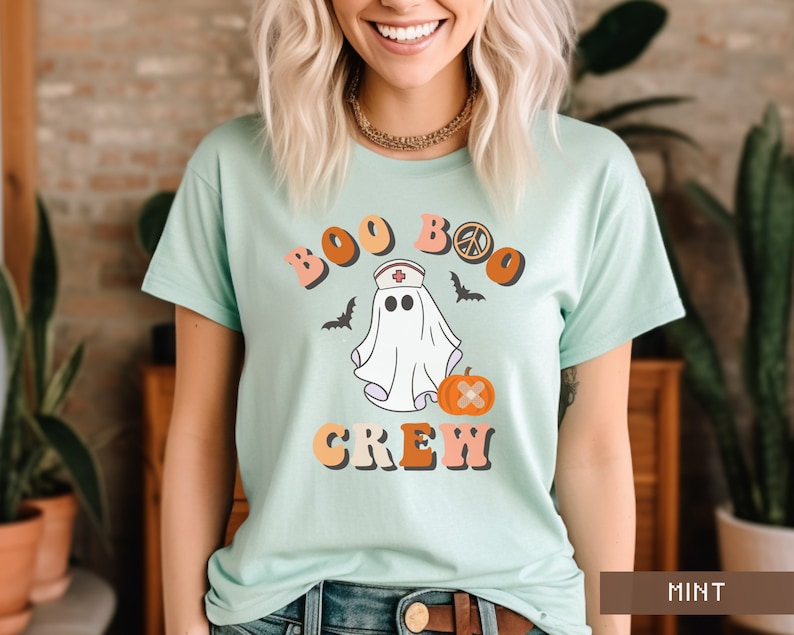 May include: A light blue t-shirt with a ghost wearing a nurse's hat and the text "Boo Boo Crew" in a retro font. The ghost is holding a pumpkin with a bandage on it.