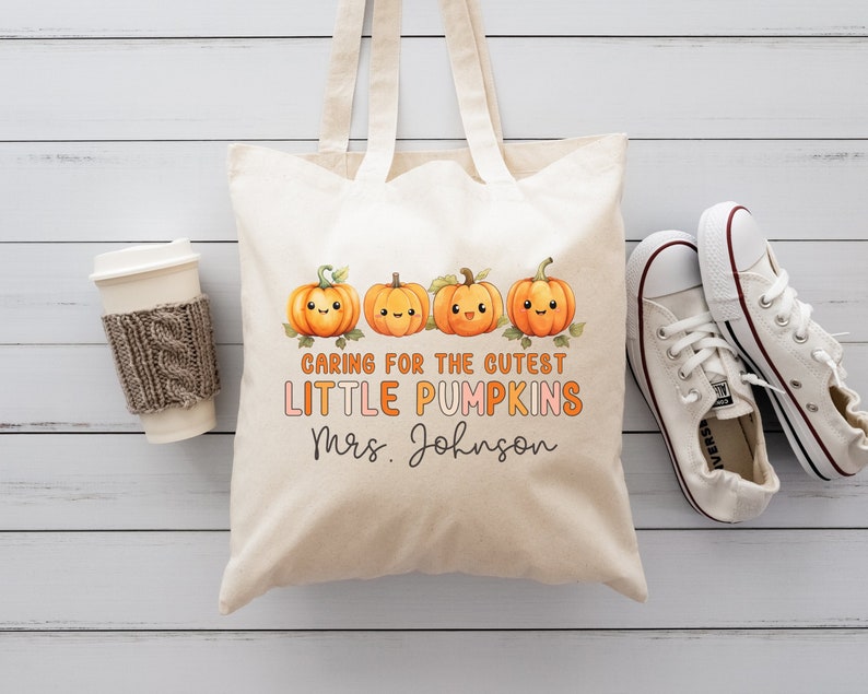 K&ouml;nnte beinhalten: Eine wei&szlig;e Stofftasche mit einem Design aus vier orangefarbenen K&uuml;rbissen mit l&auml;chelnden Gesichtern. Der Text auf der Tasche lautet "Caring for the cutest little pumpkins Mrs. Johnson".