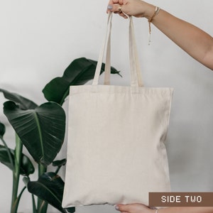 K&ouml;nnte beinhalten: Eine schlichte wei&szlig;e Canvas-Tragetasche mit zwei Henkeln. Die Tasche wird von einer Person in der Hand gehalten. Der Text "SIDE TWO" ist auf einem braunen Etikett am unteren Rand des Bildes aufgedruckt.