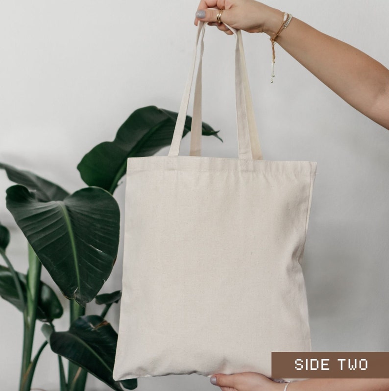 K&ouml;nnte beinhalten: Eine schlichte wei&szlig;e Canvas-Tragetasche mit zwei Henkeln. Die Tasche wird von einer Person in der Hand gehalten. Der Text "SIDE TWO" ist auf einem braunen Etikett am unteren Rand des Bildes aufgedruckt.