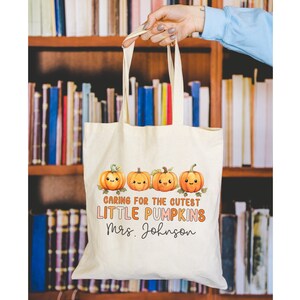 K&ouml;nnte beinhalten: Eine wei&szlig;e Stofftasche mit einem Design aus f&uuml;nf orangefarbenen K&uuml;rbissen mit l&auml;chelnden Gesichtern. Der Text auf der Tasche lautet "Caring for the cutest little pumpkins Mrs. Johnson".